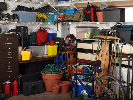 A cluttered garage interior filled with various items including plastic storage bins, cardboard boxes, and toolboxes on metal shelves at the top and on the floor. The shelves hold a mix of black, grey, and blue containers, with some yellow and red items among them. On the left side, there are black and grey filing cabinets, alongside a small yellow plastic container and a red bag. In the center, a large terracotta pot is surrounded by smaller pots, with a green garden hose coiled and placed nearby. To the right, a blue bicycle leans against the shelves, partially obscuring a wooden board and an extension cord hanging from the shelves. Scattered around are cleaning supplies, a portable radio, and miscellaneous household items, all set in a typical storage area used for rubbish and clutter clearances, likely in a residential property near Wood Green, London, associated with the Company Office Clearance Wood Green that provides rubbish removal services in the area.