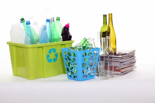 A collection of glass and plastic bottles, some with green, blue, and clear glass, are placed on a plain white surface, with a pile of newspapers or magazines beside them. The bottles are arranged in a green recycling bin with a blue plastic basket nearby, both designed for separating recyclable waste. The background is plain and neutral, emphasizing the waste items, which include various drink containers like wine bottles, beer bottles, and water bottles, some of which are partially filled or empty. The green and blue containers are made of plastic with a glossy finish, and the newspapers are stacked in an orderly pile, suggesting a typical recycling or rubbish collection setup. This scene is reflective of environmentally conscious waste separation practices, relevant to rubbish removal services offered by Office Clearance Woodgreen, located near postcode N22 in Woodgreen, London. The arrangement highlights the need for proper disposal and collection of recyclable waste as part of regular office clearance or rubbish removal processes on the local high street or commercial areas.